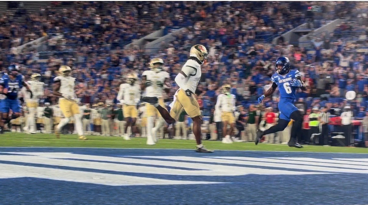 A Memphis player in a blue jersey crosses the goal line as UAB defenders trail behind him