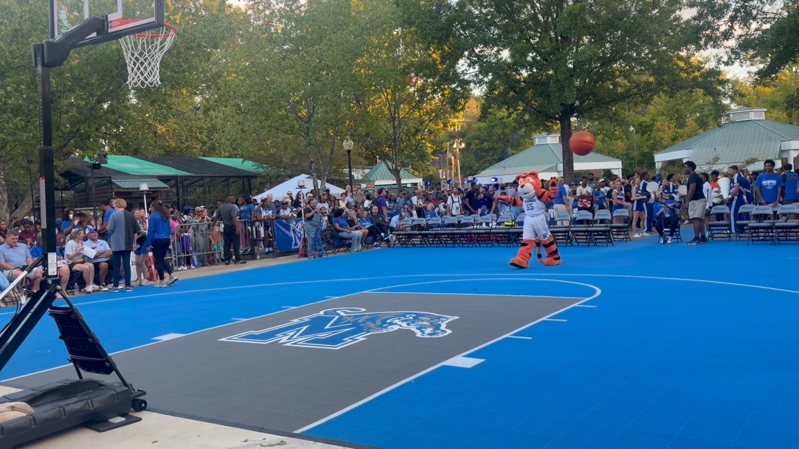 An outdoor blue basketball court and hoop ringed by trees and seated spectators